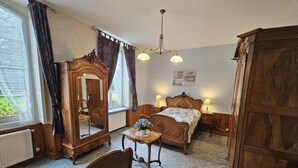 A traditional bedroom with a wooden bed, a nightstand with a lamp, a dresser, a mirror, and a window with curtains. - Gîte Du Theil Au Servant (LA CHAPELLE-VIVIERS)