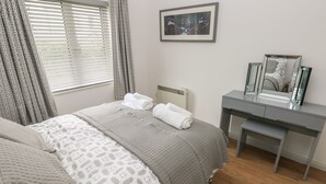 Un dormitorio con cama, una mesita de noche con espejo y una ventana con cortinas.
