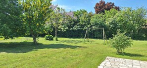 A grassy outdoor area with a swing set, trees, and a paved walkway.
