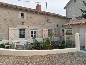 A rustic stone building with a tiled roof, a small window, and a door. There is a white table with chairs and a garden with flowers and a metal plant stand. - Le Gîte du Porche - Saint-georges-les-baillargeaux (SAINT-GEORGES-LES-BAILLARGEAUX)