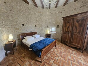 Quarto com parede de pedra, cama de madeira, mesa de cabeceira com abajur, guarda-roupa e padrão de piso em padrão herringbone.