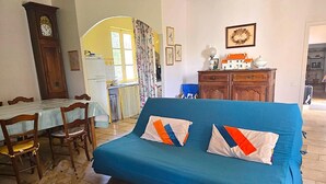 Sala de estar con sofá azul, mesa de comedor de madera, sillas, un reloj y cocina visible a través de un arco.