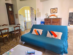Un salon avec un canapé bleu, une table à manger en bois, des chaises, une horloge et une cuisine visible à travers une arche.