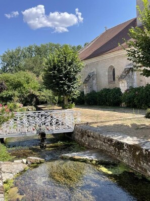 A historic building with a stone facade, a white decorative bridge over a small stream, and lush greenery in the background. - Le Petit Ru - Lizant (LIZANT)