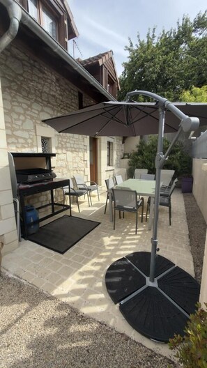 Un patio al aire libre con una parrilla, mesa, sillas y un paraguas grande.