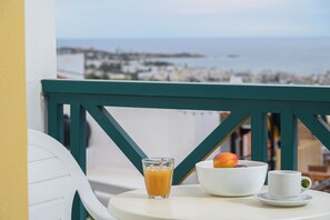 A table with a glass of orange juice, a bowl of fruit, and a cup on a balcony overlooking a coastal city. - Elen-Mari (Hersonissos)