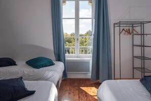 Ein Zimmer mit zwei Betten, einem Fenster mit Vorhängen, einem Heizkörper und einem Metallregal.