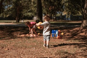 Ein Kind spielt mit einem Ball in einem Park mit Bäumen und einem Spielplatz im Hintergrund.