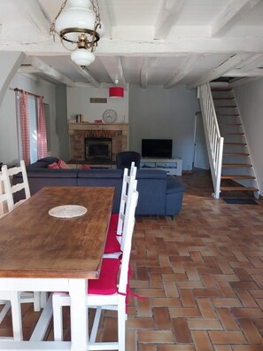 Un comedor con una mesa de madera y sillas blancas, un sofá, una chimenea, un televisor y una escalera.