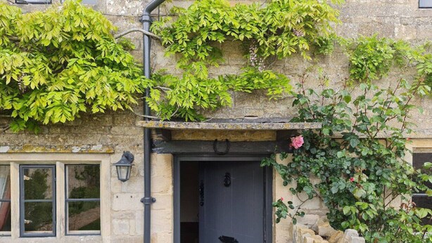 A stone building with a dark door, a small window, and a stone wall with plants. - Charming cottage in the heart of the Cotswolds (Cirencester)