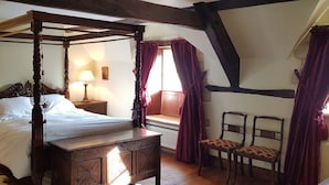 A four-poster bed with a wooden headboard, a nightstand with a lamp, a window with curtains, and a chair with a patterned cushion. - Charming cottage in the heart of the Cotswolds (Cirencester)