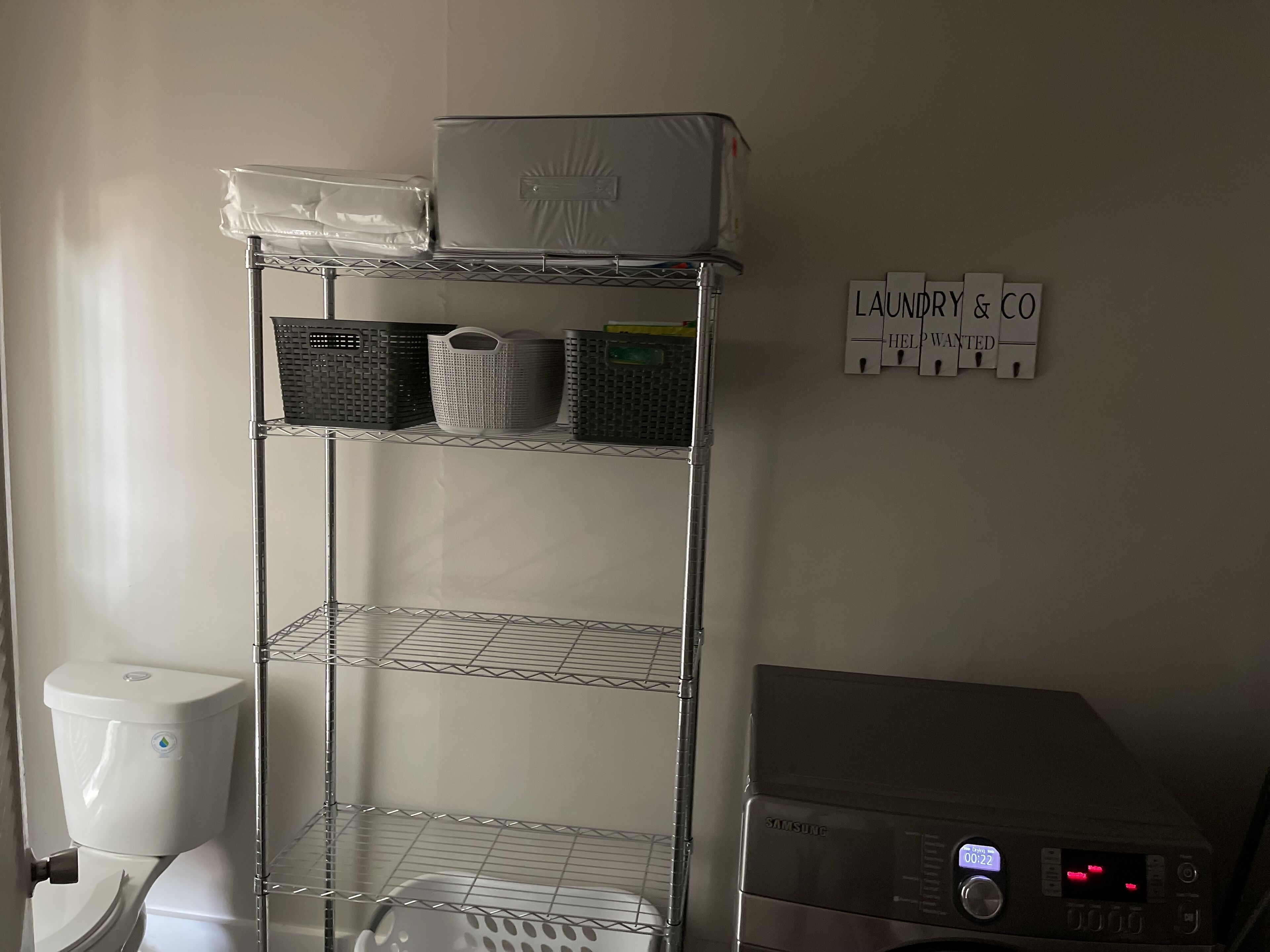 Een was- en verzorgingsstation met een Samsung wasmachine, een metalen rek met manden, en een toilet.