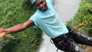 Una persona saltando en bungee con los brazos extendidos, con casco y arnés, frente a un exuberante valle verde y una cascada.
