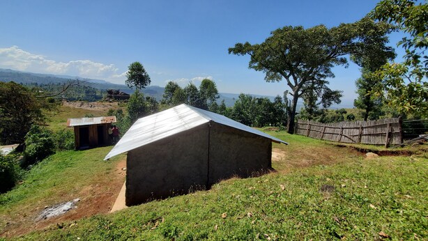 Un entorno rural con un pequeño edificio de una sola planta con techo de metal, rodeado de vegetación y una valla de madera.