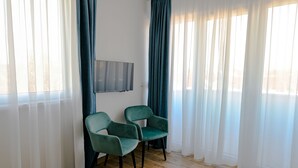 A room with two teal chairs, a flat-screen TV mounted on the wall, and a large window with sheer white curtains and dark blue drapes. - Dona (Belgrade)