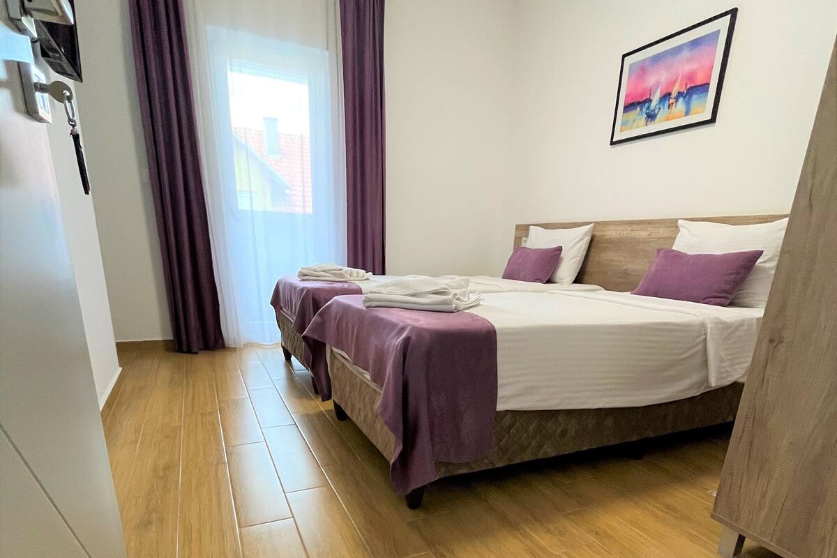 A bedroom with a bed, a window with curtains, a painting on the wall, and a wooden floor.