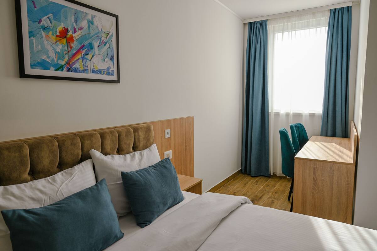 A hotel room with a bed, a desk, and a chair. There is a painting on the wall and a window with curtains.