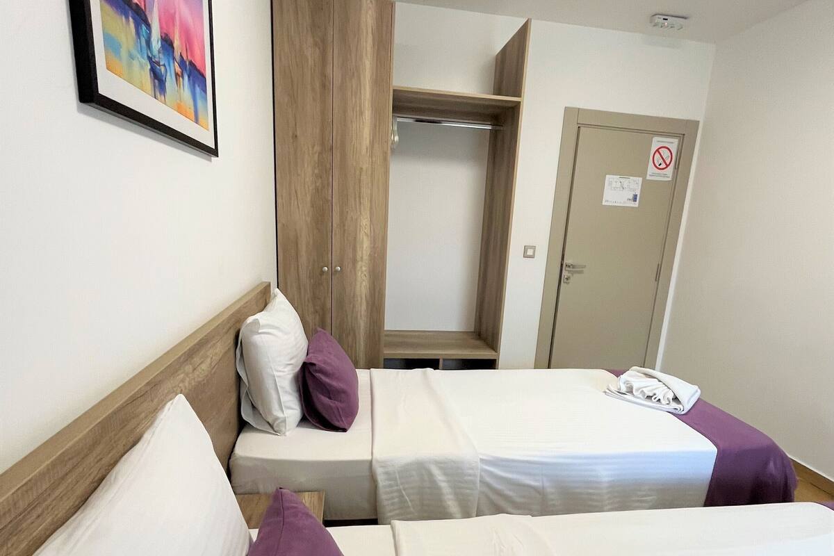 A hotel room with two beds, a wooden wardrobe, a door with a no-smoking sign, and a framed picture on the wall.