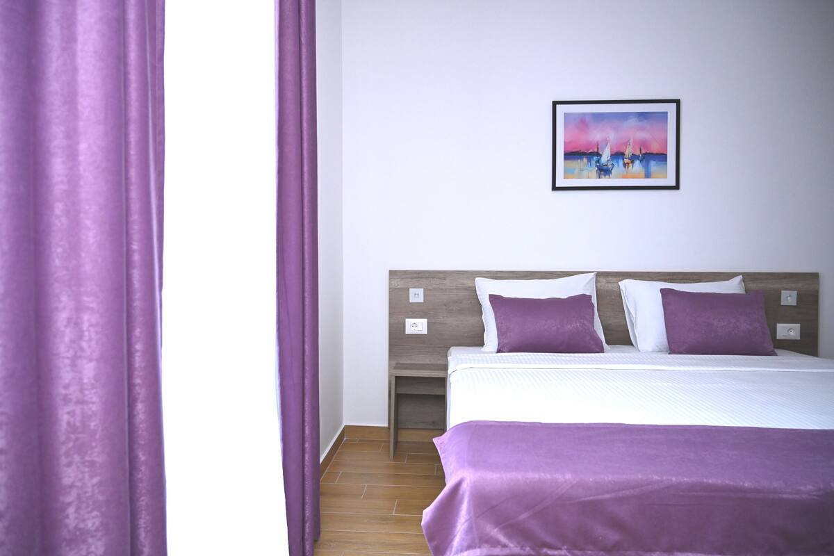 A hotel room with a bed, purple bedding, a nightstand, a wall-mounted painting, and a wooden floor.