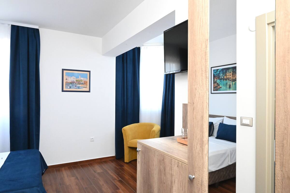 A hotel room with a bed, a wooden cabinet, a yellow armchair, and two paintings on the walls.