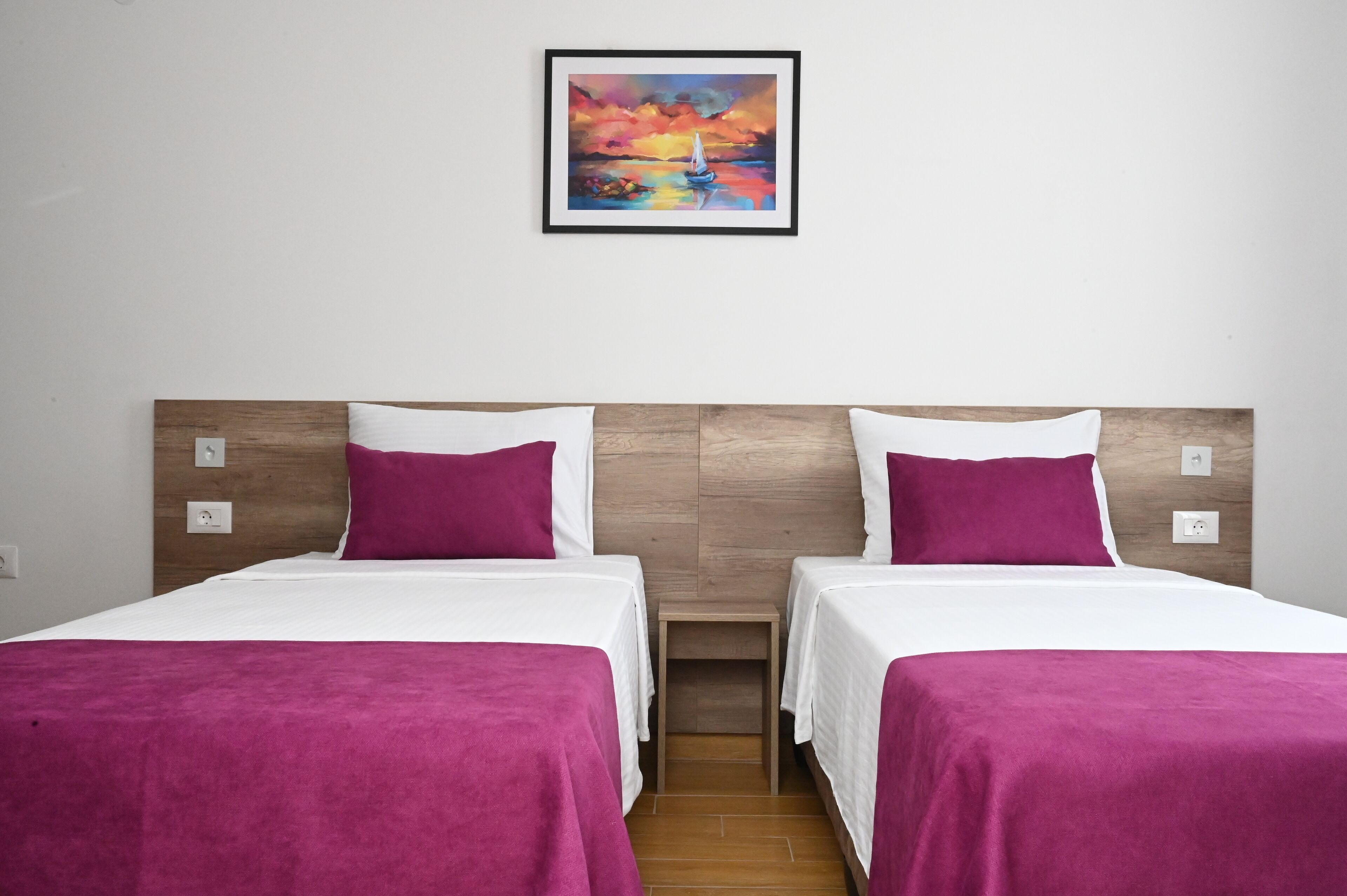 Een tweepersoonskamer met twee bedden, elk met een houten hoofdbord, wit beddengoed en een paars plaid. Boven de bedden hangt een lijst met een schilderij van een zonsondergang over water.