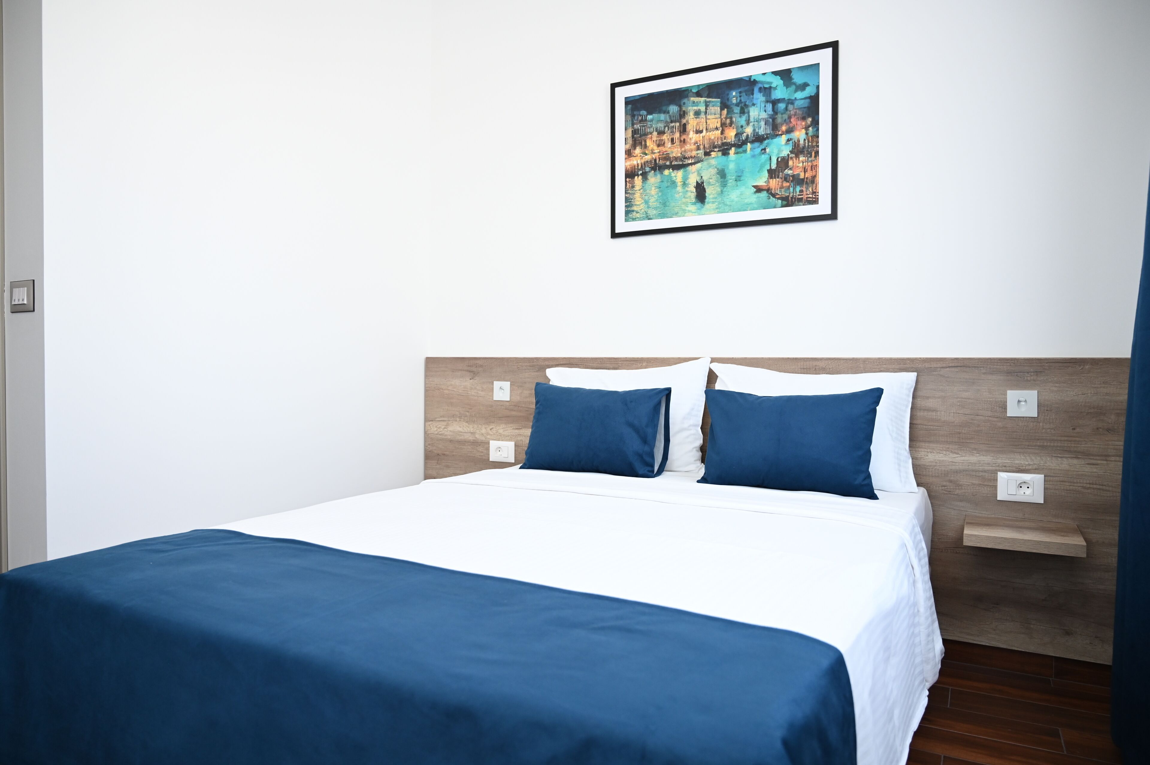 A neatly made bed with a blue bedspread and pillows, a wooden headboard, and a framed painting of a cityscape on the wall.