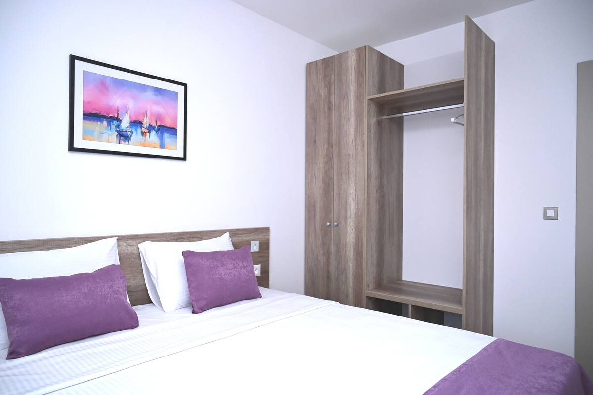 A bedroom with a bed, two purple pillows, a wooden wardrobe, and a framed painting on the wall.
