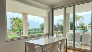 A modern dining area with a glass table and white chairs, large windows with a view of greenery and a pool, and a sliding glass door leading to an outdoor patio.