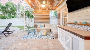 A covered outdoor patio with a wooden ceiling, a sofa, a TV, and a bar area.