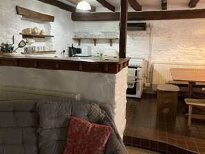 A cozy kitchen with white walls and wooden beams, a dining area with a wooden table and chairs, and a grey sofa with a red pillow. - Relax in Mountain View Cottage at Cwmiar Farm (Llanybydder)