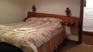 Un dormitorio con cabecera de madera, ropa de cama estampada con motivos florales, una mesita de noche con lámpara y un pequeño adorno en la pared.