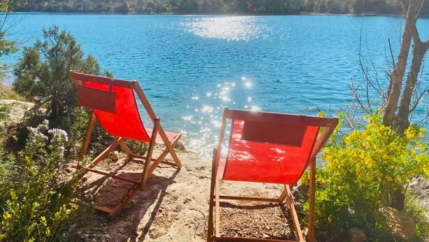 Two red folding chairs on a sandy beach overlooking a lake with a forested hill in the background.
