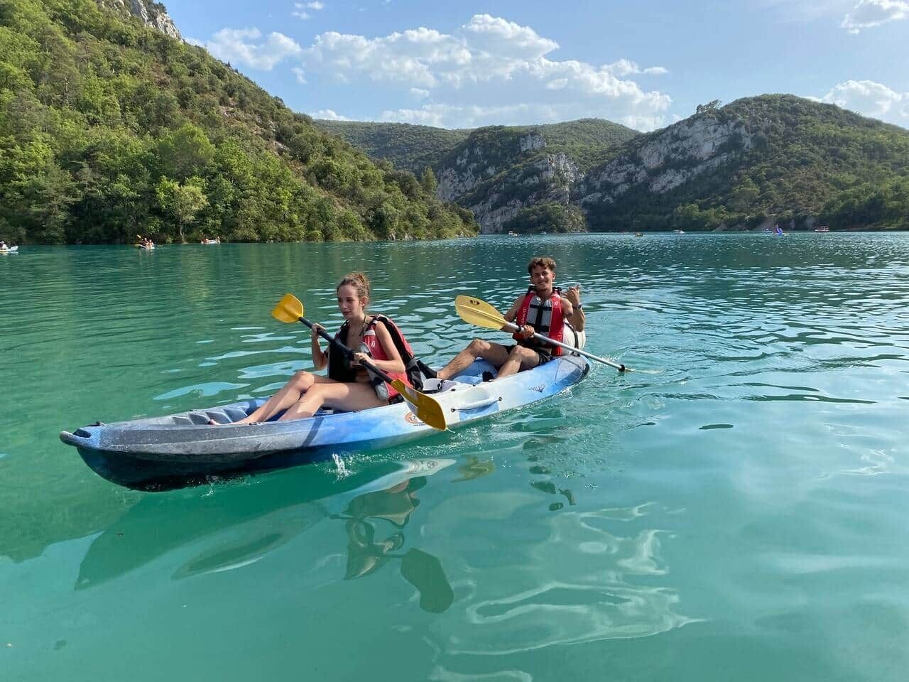 Two people in a kayak on a calm lake with mountains in the background.