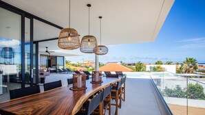 A modern dining area with a wooden table and chairs, hanging wicker light fixtures, and a view of the ocean and palm trees.
