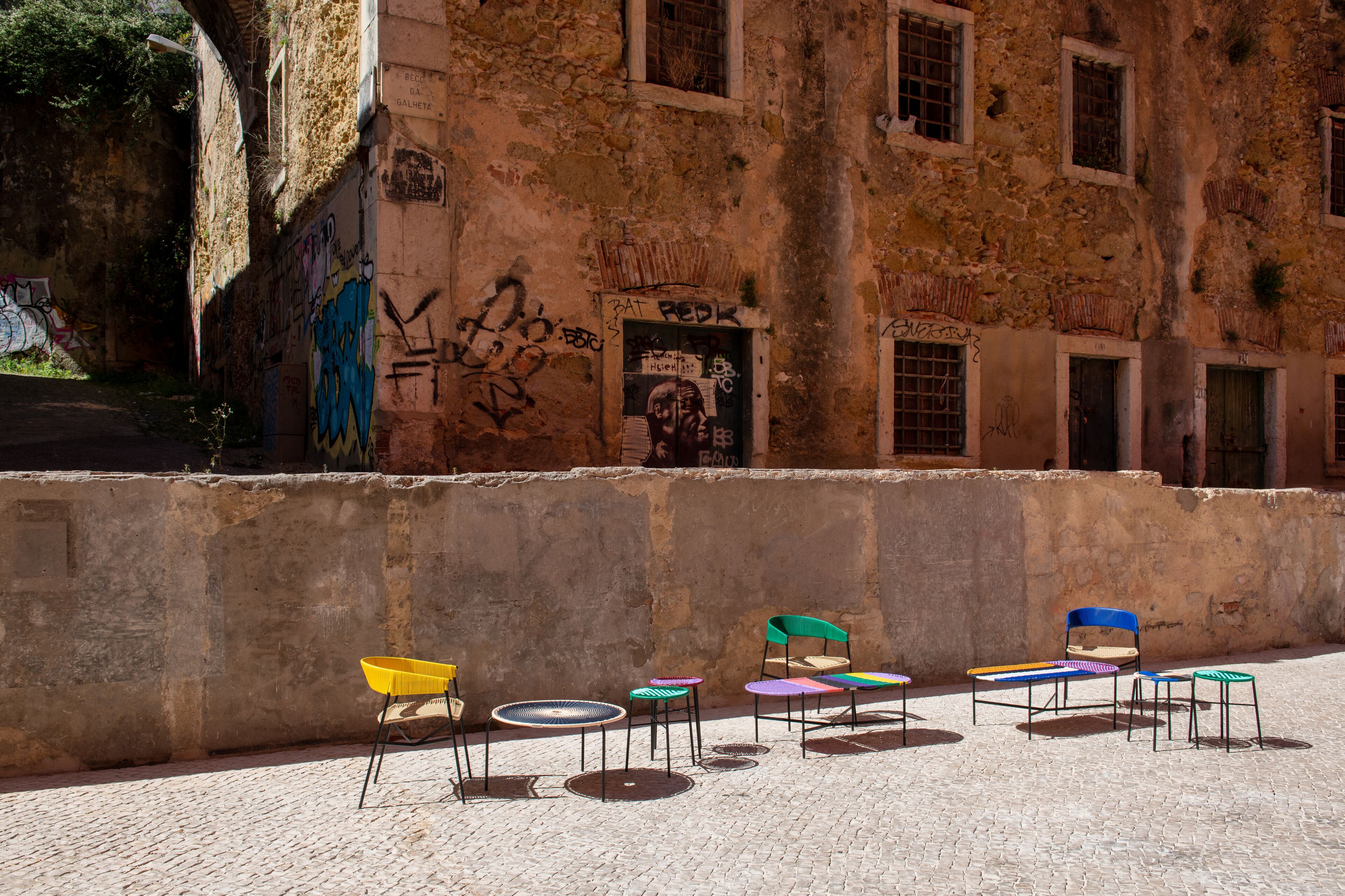 A courtyard with a row of colorful chairs and tables, a stone wall with graffiti, and a building with multiple windows and a door.