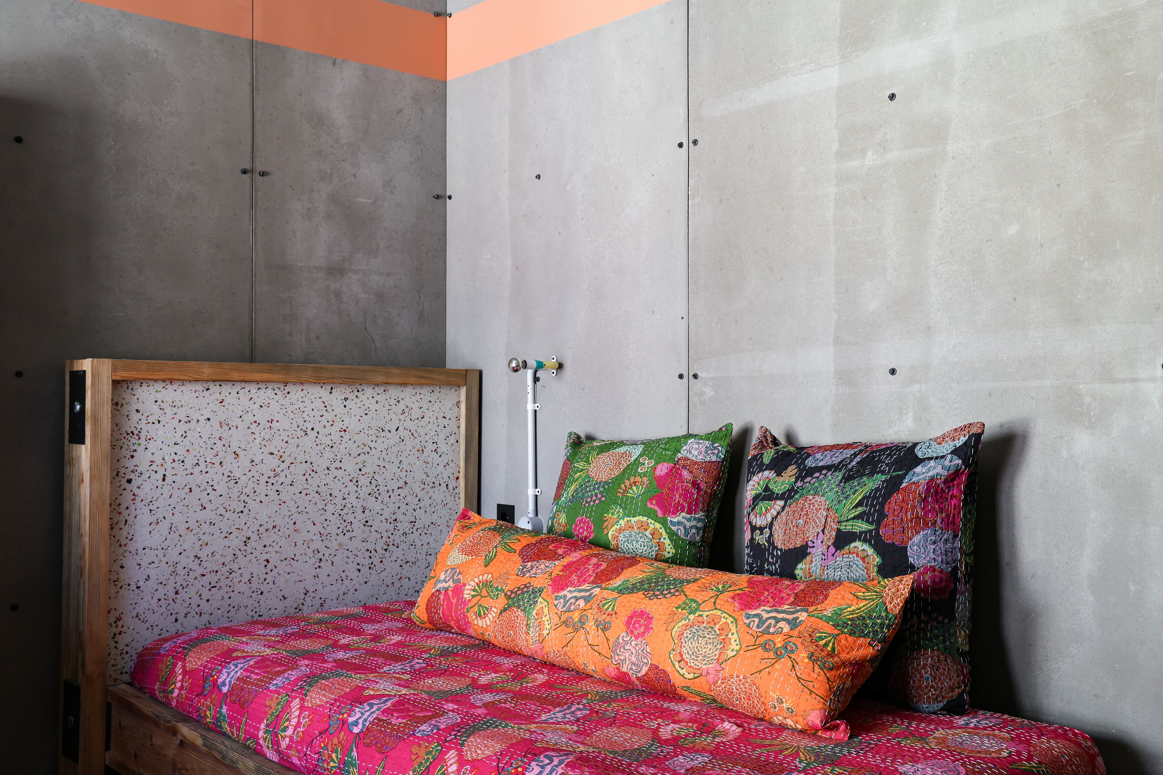 Una cama con estructura de madera y ropa de cama floral colorida en una habitación con paredes de concreto y un diseño sencillo y moderno.