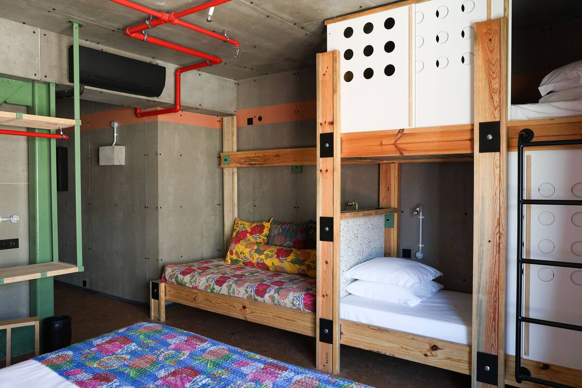 A bunk bed room with a wooden bunk bed, a lower bunk with a colorful quilt, a small wooden shelf, and a bunk bed with a white mattress.