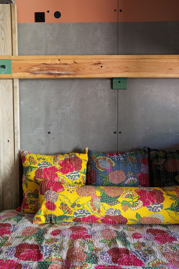 A bed with floral-patterned pillows and a quilt, a wooden headboard, and a grey wall with a door and two switches. - Jam Hotel Lisbon (Lisbon)
