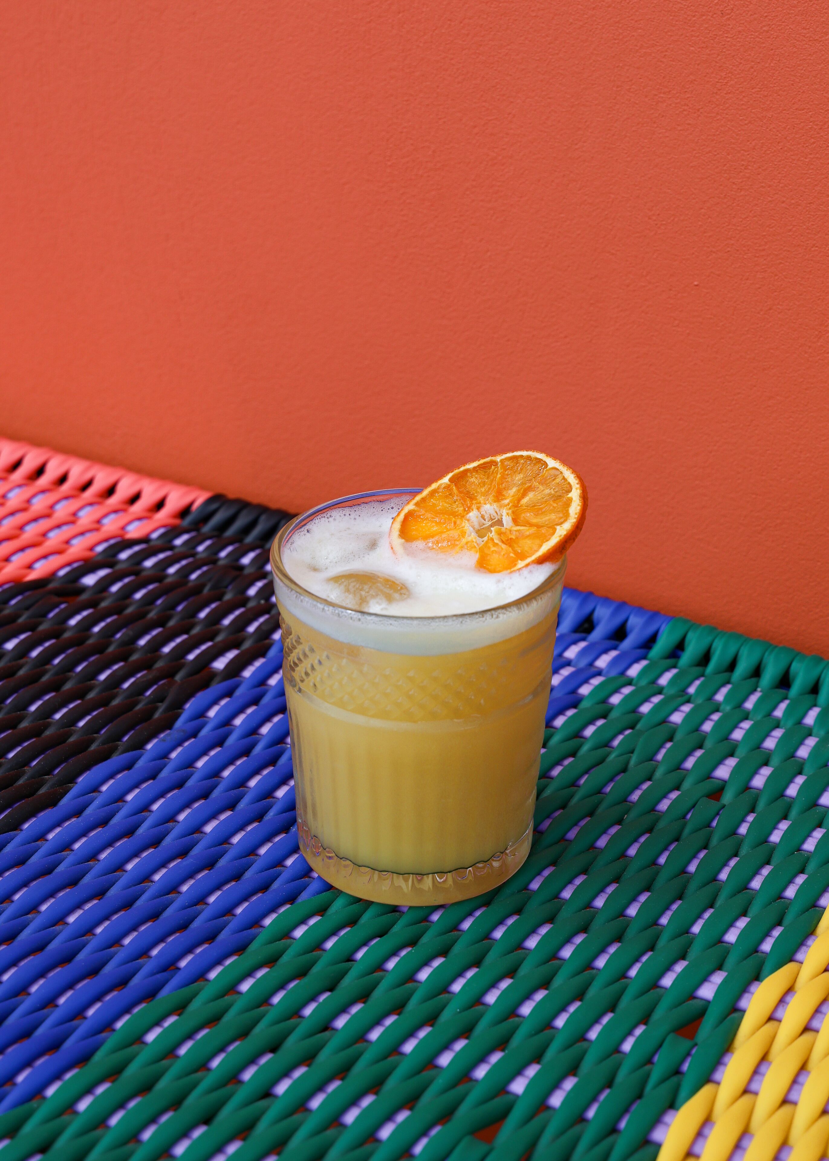 A glass of orange juice with a slice of orange on a woven table against a red wall.