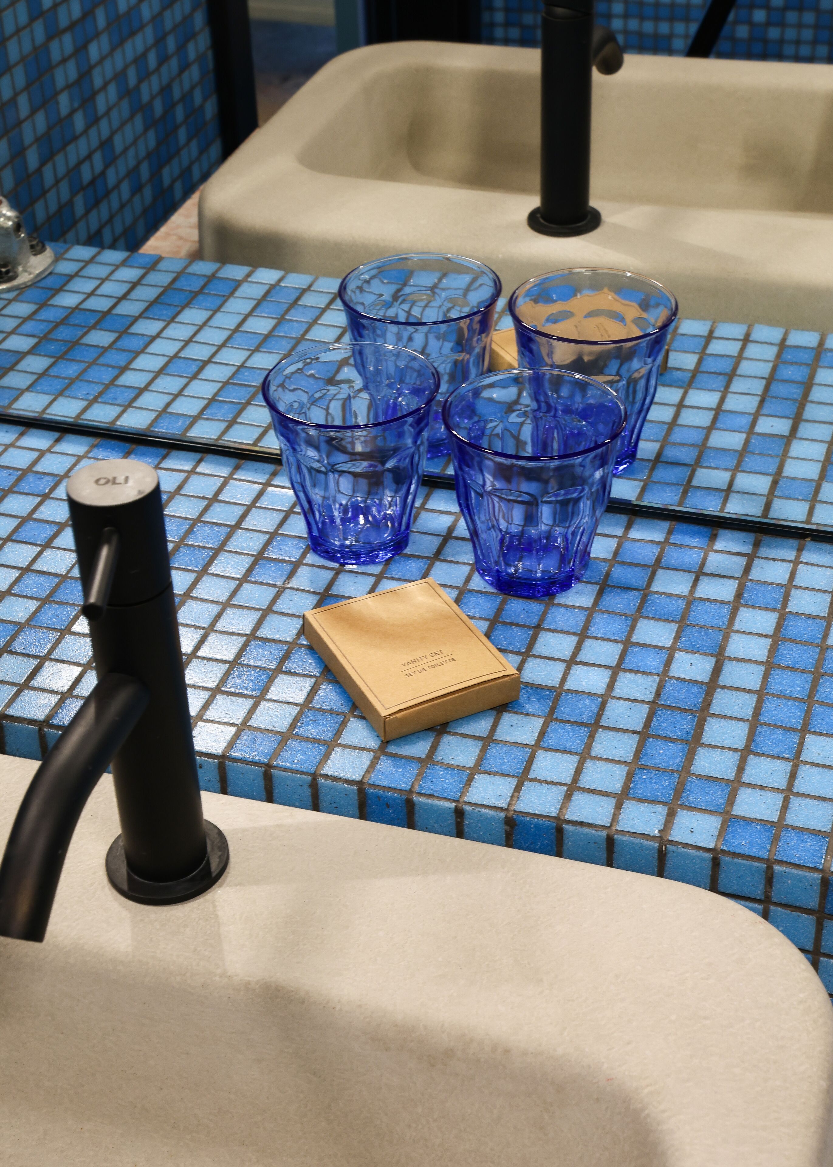 A bathroom sink with a blue tiled backsplash, four blue glasses, and a small brown box labeled "Vanity Set 507 DE TOILETTE".