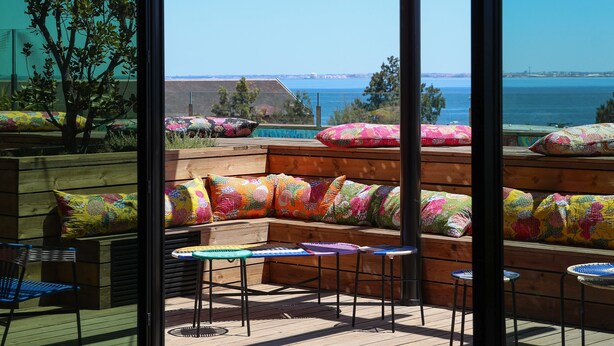 A patio with wooden benches and colorful cushions, overlooking a body of water and a distant cityscape. - Jam Hotel Lisbon (Lisbon)