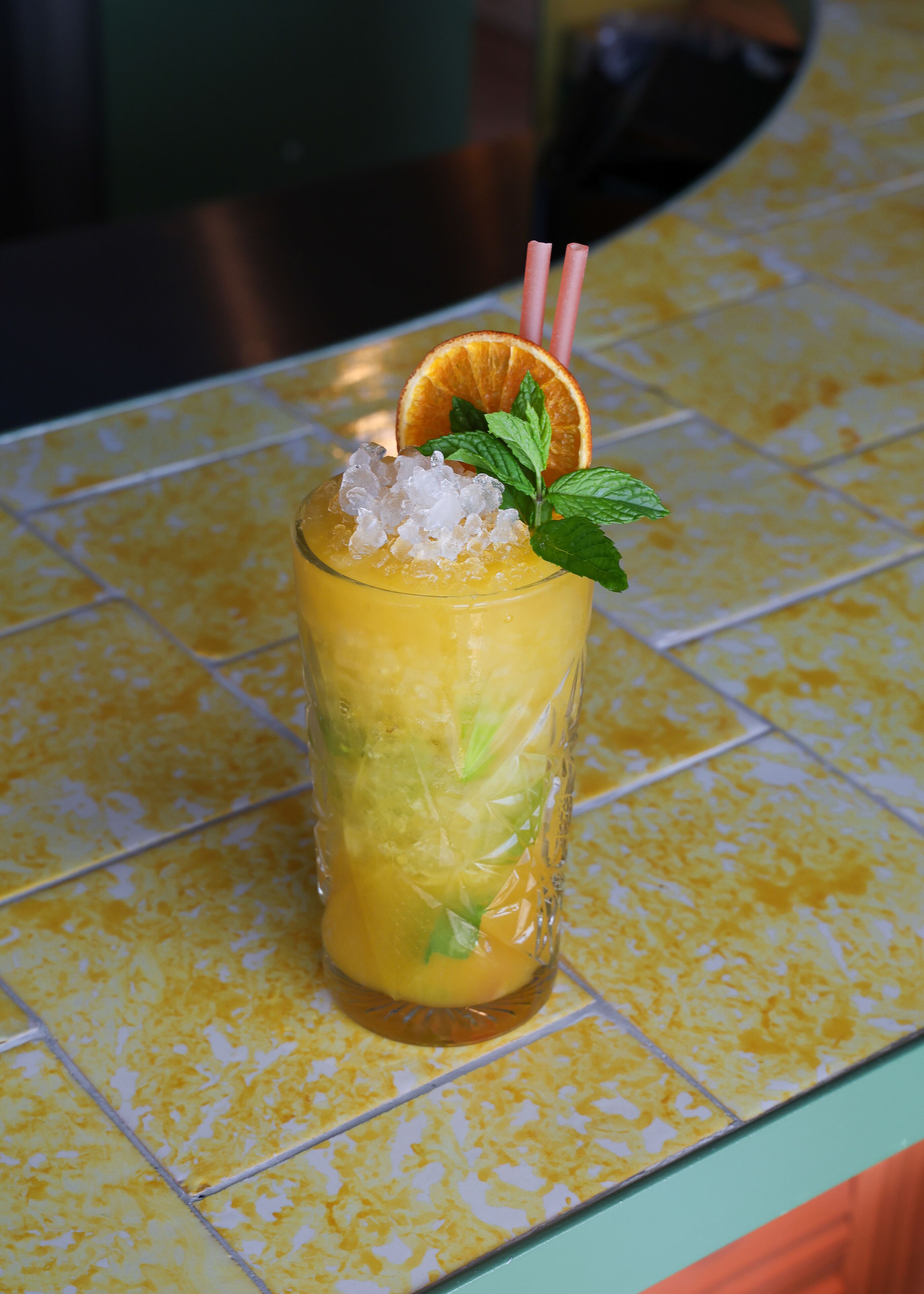 A tall glass with a yellow drink, ice cubes, a slice of orange, mint leaves, and a straw.