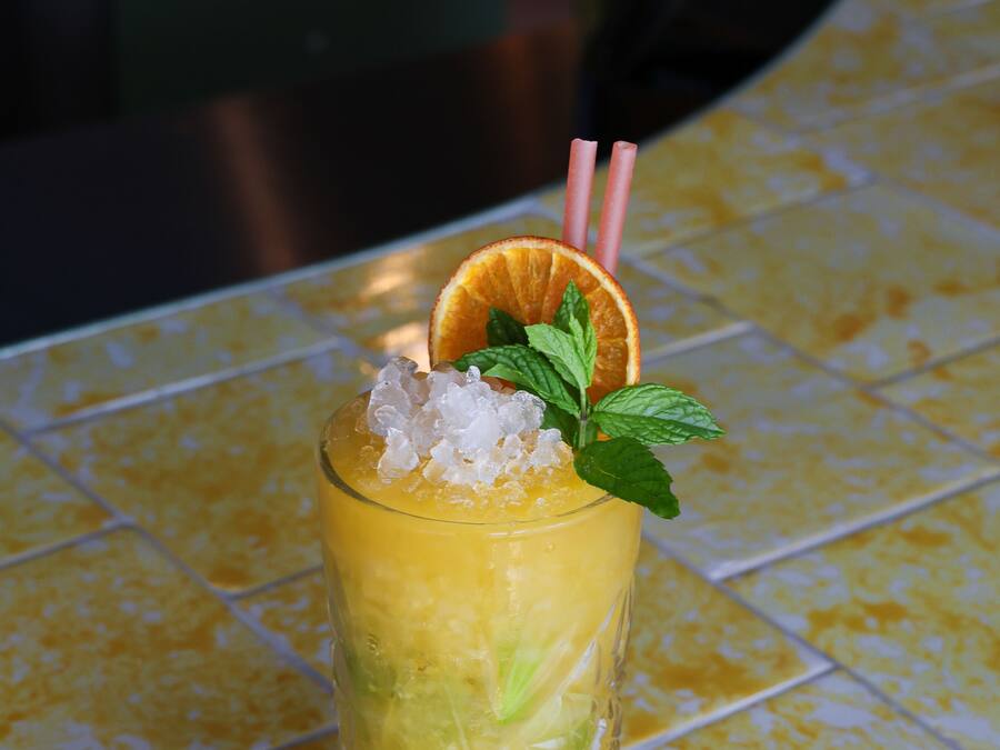 A tall glass with a yellow drink, ice cubes, a slice of orange, mint leaves, and a straw.