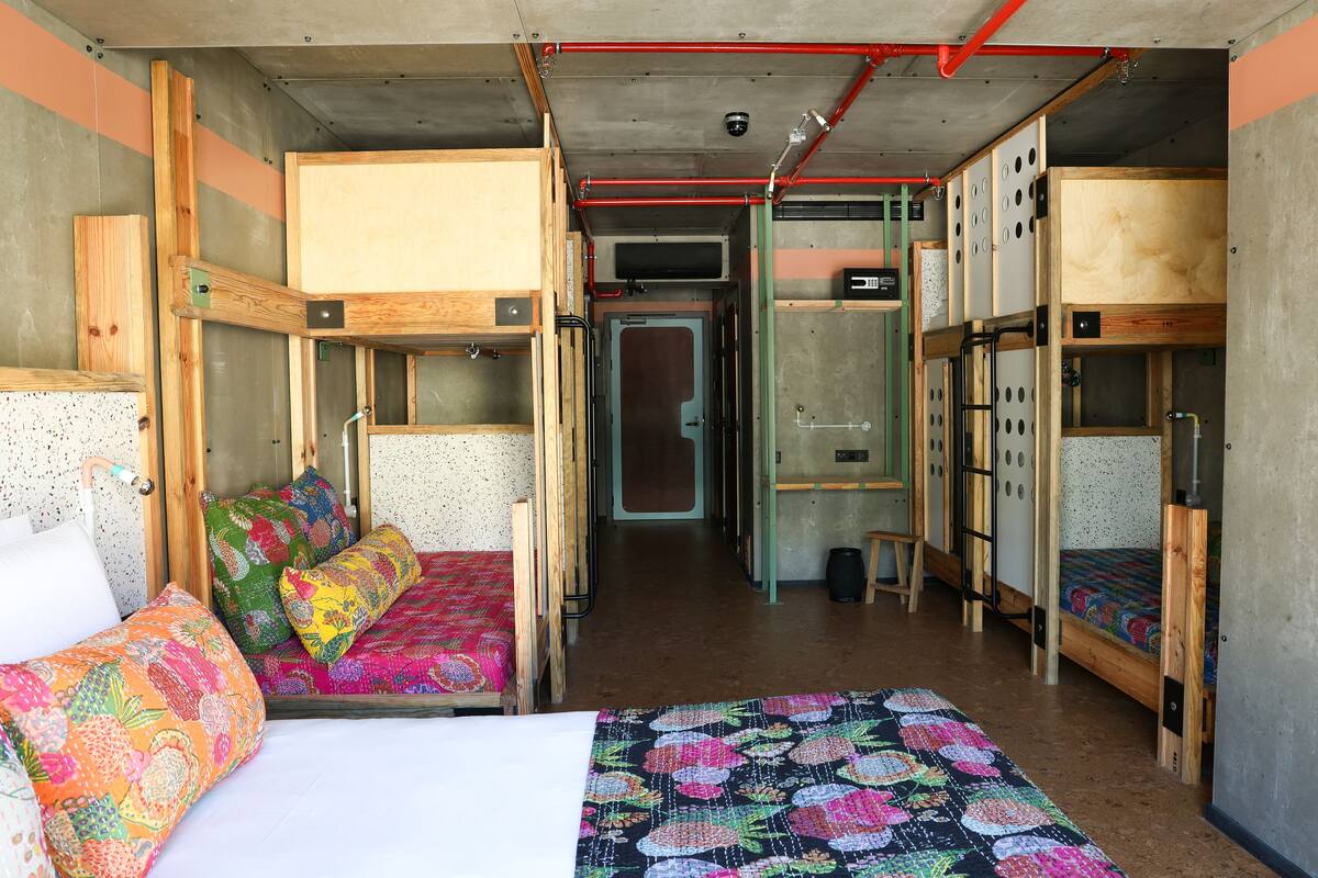 A dormitory room with bunk beds, a small kitchenette, and a door leading to another room.