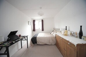 Ein Schlafzimmer mit Bett, Schreibtisch mit Laptop, Nachttisch mit Pflegeprodukten und einem Fenster mit Vorhängen.