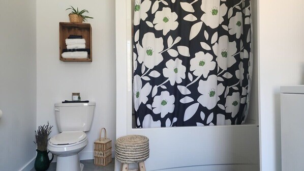 A bathroom with a toilet, a shower curtain with a floral pattern, a small wooden shelf with towels, and a wicker basket. - Le Bétula - Beach and lake view (Saint-Félicien)
