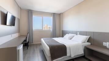 Quarto de hotel moderno com cama, mesa de cabeceira, televisão e janela com vista para prédios.