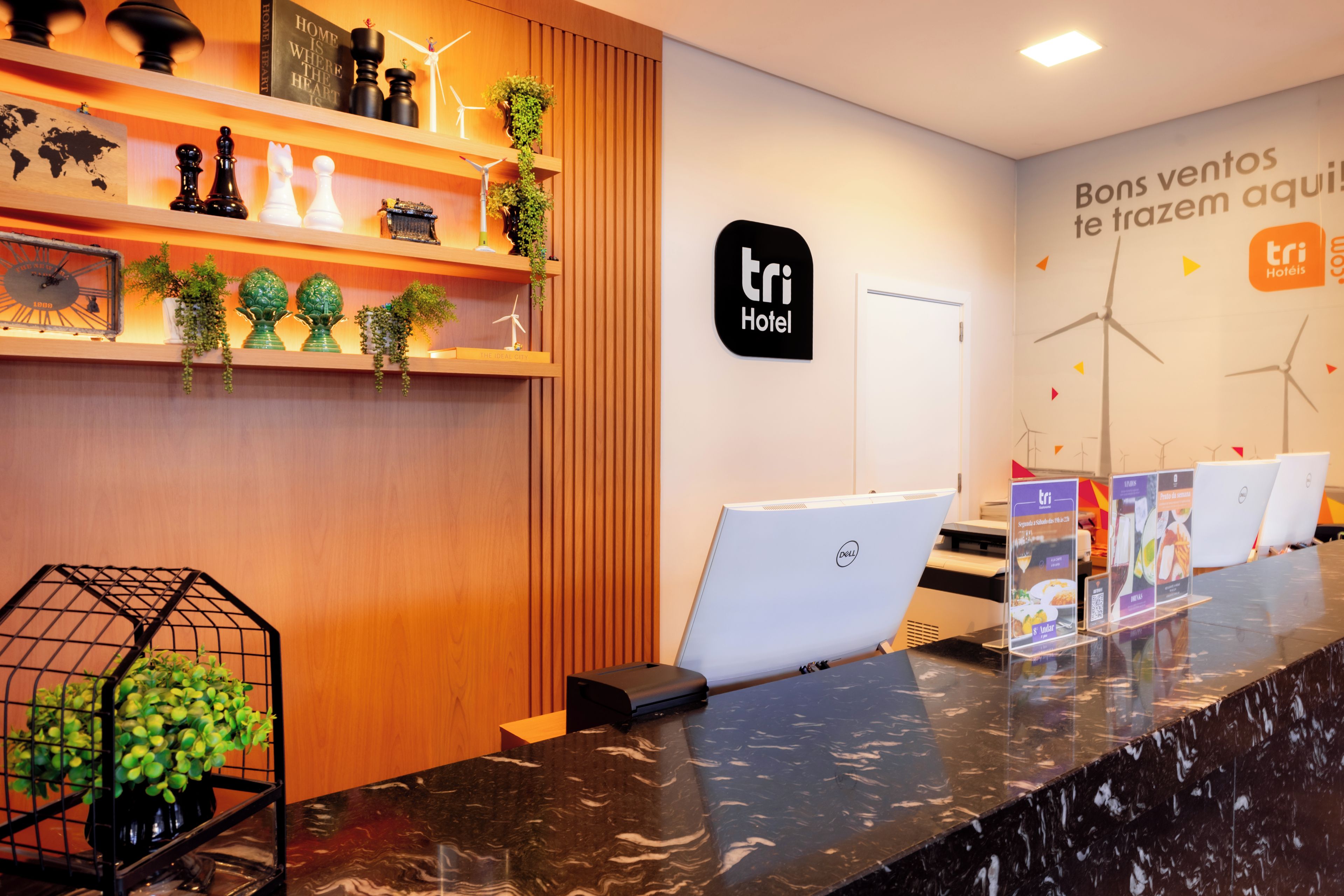 A hotel reception desk with a wooden panel wall, a black sign with 'tri Hotel' logo, and wind turbines on the wall.