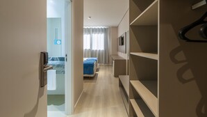 Habitación de hotel con piso de madera, cama con cubrecama azul, armario y baño con puerta de cristal.
