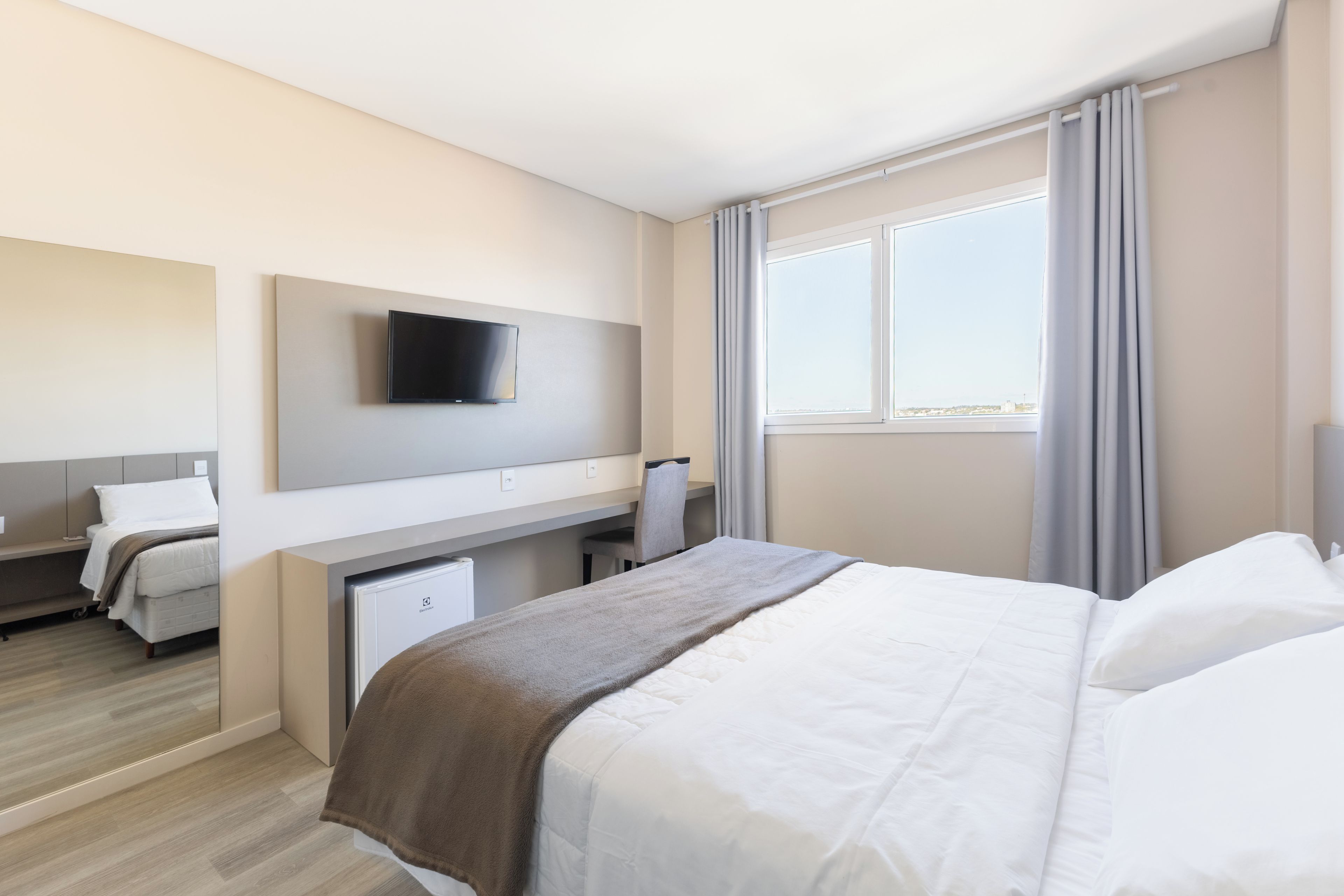 Habitación de hotel con cama, escritorio, silla, televisor montado en la pared y ventana con cortinas.